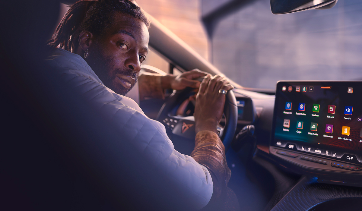 Fahrer sitzt am Steuer eines CUPRA Modells, Blick in die Kamera, im Vordergrund das große Infotainment-Display mit farbigen App-Icons